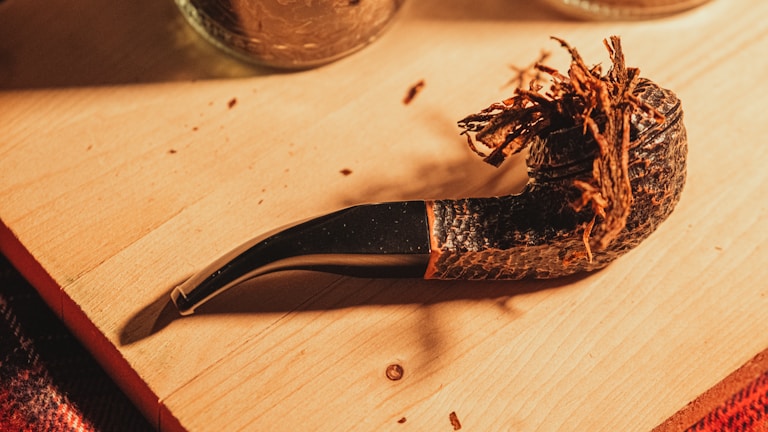 A wooden pipe with dried tobacco strands is placed on a wooden surface. The pipe has a textured, dark brown finish and a curved stem. Nearby, parts of glass jars are visible, suggesting a cozy setting.