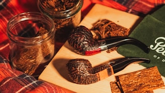 Two rustic smoking pipes are placed alongside glass jars filled with tobacco on a wooden board. A green cloth with cursive lettering is partially visible, and a plaid fabric background adds warmth to the scene.