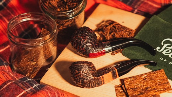 Two rustic smoking pipes are placed alongside glass jars filled with tobacco on a wooden board. A green cloth with cursive lettering is partially visible, and a plaid fabric background adds warmth to the scene.