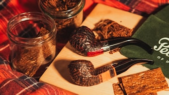 Two rustic smoking pipes are placed alongside glass jars filled with tobacco on a wooden board. A green cloth with cursive lettering is partially visible, and a plaid fabric background adds warmth to the scene.