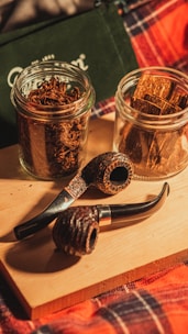 Two jars containing tobacco sit on a wooden board alongside two smoking pipes. The surrounding is cozy, featuring a plaid fabric with red and blue tones, suggesting a warm, rustic atmosphere.
