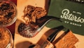 A collection of smoking pipes and tobacco is arranged on a wooden surface. The pipes have dark, textured bowls and smooth, black stems. There are also jars of loose tobacco and pieces of pressed tobacco. A green box or booklet with the name 'Peterson of Dublin' and an establishment date in white lettering is visible.