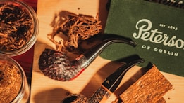 A collection of smoking pipes and tobacco is arranged on a wooden surface. The pipes have dark, textured bowls and smooth, black stems. There are also jars of loose tobacco and pieces of pressed tobacco. A green box or booklet with the name 'Peterson of Dublin' and an establishment date in white lettering is visible.