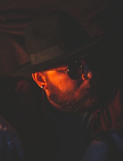A person with a beard is wearing sunglasses and a wide-brimmed hat. The lighting is low and warm, casting strong shadows across the face, creating a mysterious and contemplative atmosphere.