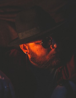 A person with a beard is wearing sunglasses and a wide-brimmed hat. The lighting is low and warm, casting strong shadows across the face, creating a mysterious and contemplative atmosphere.