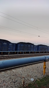 A train consisting of multiple freight cars is parked on railway tracks. Each car appears to be filled with coal. Overhead power lines stretch across the sky, and the setting sun casts a subtle light.