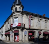 A vintage-style building with a rounded corner turret and intricate architectural details. It features red signage for a pizza restaurant that operates 24/7. The structure is made of white stone with a dark gray roof, and there are multiple arched windows with closed shutters.