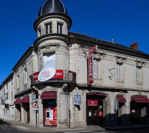 A vintage-style building with a rounded corner turret and intricate architectural details. It features red signage for a pizza restaurant that operates 24/7. The structure is made of white stone with a dark gray roof, and there are multiple arched windows with closed shutters.