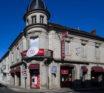 A vintage-style building with a rounded corner turret and intricate architectural details. It features red signage for a pizza restaurant that operates 24/7. The structure is made of white stone with a dark gray roof, and there are multiple arched windows with closed shutters.
