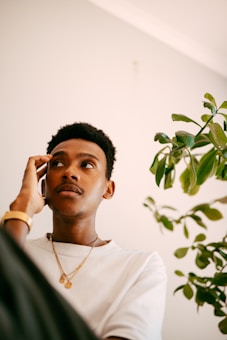 A person is engaged in a phone call, appearing thoughtful. Wearing a casual white t-shirt and gold jewelry, the individual is sitting indoors next to a leafy green plant. Sunlight softly illuminates the space, creating a warm, calm atmosphere.