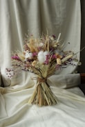 A bouquet of dried flowers arranged with soft, muted colors. The composition includes various textures and shades, featuring clusters of purple, beige, and white blossoms. The bouquet is tied with a simple natural string and is set against a creamy fabric background, which adds a warm and rustic ambiance.