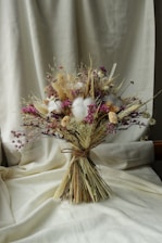 A bouquet of dried flowers arranged with soft, muted colors. The composition includes various textures and shades, featuring clusters of purple, beige, and white blossoms. The bouquet is tied with a simple natural string and is set against a creamy fabric background, which adds a warm and rustic ambiance.