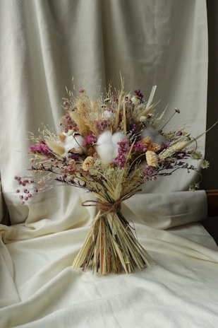 A bouquet of dried flowers arranged with soft, muted colors. The composition includes various textures and shades, featuring clusters of purple, beige, and white blossoms. The bouquet is tied with a simple natural string and is set against a creamy fabric background, which adds a warm and rustic ambiance.