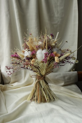 A bouquet of dried flowers arranged with soft, muted colors. The composition includes various textures and shades, featuring clusters of purple, beige, and white blossoms. The bouquet is tied with a simple natural string and is set against a creamy fabric background, which adds a warm and rustic ambiance.