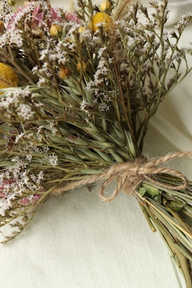 A rustic bouquet of dried flowers tied together with twine lies on a light-colored fabric surface. The bouquet includes a mix of small white blossoms, yellow round flowers, and a few sprigs of grasses and green leaves.