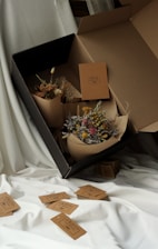 A large cardboard box positioned on white fabric contains two bouquets of dried flowers. The flowers are wrapped in simple brown paper. Several brown tags with text are scattered around on the fabric, with one visible inside the box.