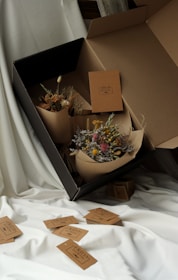 A large cardboard box positioned on white fabric contains two bouquets of dried flowers. The flowers are wrapped in simple brown paper. Several brown tags with text are scattered around on the fabric, with one visible inside the box.