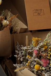 A collection of dried floral arrangements in brown paper wrapping, featuring small white flowers, bright yellow and pink blooms, and various grasses. A cardboard box with Cyrillic script is positioned in the background.