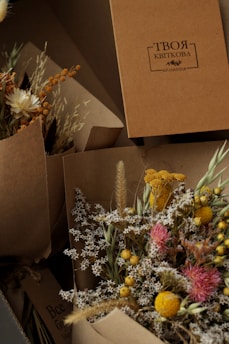 A collection of dried floral arrangements in brown paper wrapping, featuring small white flowers, bright yellow and pink blooms, and various grasses. A cardboard box with Cyrillic script is positioned in the background.