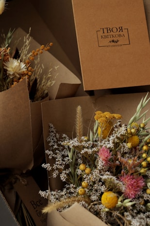 A collection of dried floral arrangements in brown paper wrapping, featuring small white flowers, bright yellow and pink blooms, and various grasses. A cardboard box with Cyrillic script is positioned in the background.