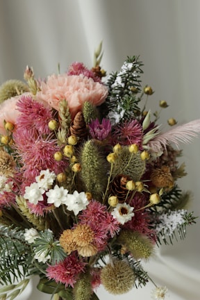 An arrangement of dried flowers and plants with various textures and colors, including pink, white, green, and yellow blooms. The bouquet features diverse flower shapes, some resembling thistles with spiky textures, others with soft petals like carnations. Among the foliage are coniferous twigs and fluffy grasses, creating a rustic and natural look.
