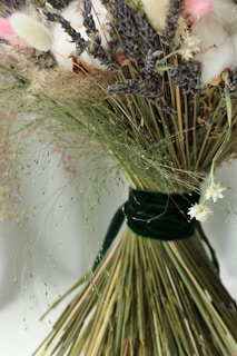 A close-up of a bouquet of various dried flowers and grasses tied together with a dark green ribbon. The mix includes delicate white, pink, and lavender-colored flowers, complemented by long, slender stems of different textures.