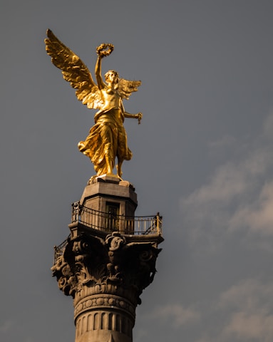 A golden angelic statue stands atop a tall, ornate column with intricate details. The figure has widespread wings and holds a laurel wreath aloft, set against a muted sky.