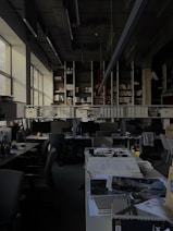 A dimly lit office space with rows of desks and chairs. Large windows on the left allow some natural light to enter. The desks are cluttered with office supplies, paperwork, and computer monitors. A long shelf above the desks holds numerous binders and books. The ceiling features exposed structural elements and suspended lighting fixtures.