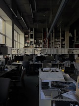 A dimly lit office space with rows of desks and chairs. Large windows on the left allow some natural light to enter. The desks are cluttered with office supplies, paperwork, and computer monitors. A long shelf above the desks holds numerous binders and books. The ceiling features exposed structural elements and suspended lighting fixtures.