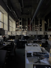 A dimly lit office space with rows of desks and chairs. Large windows on the left allow some natural light to enter. The desks are cluttered with office supplies, paperwork, and computer monitors. A long shelf above the desks holds numerous binders and books. The ceiling features exposed structural elements and suspended lighting fixtures.