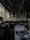 A dimly lit office space with rows of desks and chairs. Large windows on the left allow some natural light to enter. The desks are cluttered with office supplies, paperwork, and computer monitors. A long shelf above the desks holds numerous binders and books. The ceiling features exposed structural elements and suspended lighting fixtures.