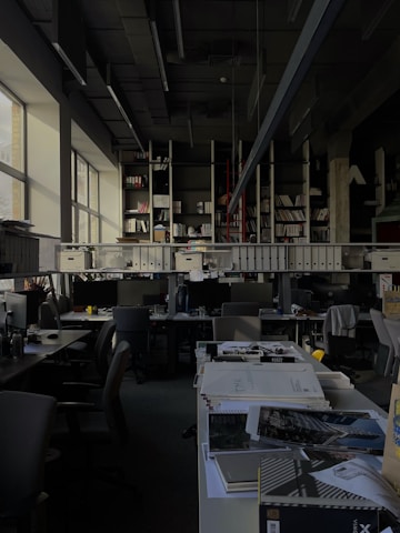 A dimly lit office space with rows of desks and chairs. Large windows on the left allow some natural light to enter. The desks are cluttered with office supplies, paperwork, and computer monitors. A long shelf above the desks holds numerous binders and books. The ceiling features exposed structural elements and suspended lighting fixtures.