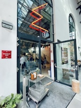 An entrance to a modern restaurant with large glass doors, featuring a red neon zigzag light above. Inside, there is a glimpse of people seated and a staircase leading to an upper level. A metal cart with two wire baskets and a hand sanitizer station is positioned by the entrance. A Michelin 2020 plaque is displayed on the left wall.