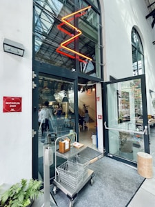 An entrance to a modern restaurant with large glass doors, featuring a red neon zigzag light above. Inside, there is a glimpse of people seated and a staircase leading to an upper level. A metal cart with two wire baskets and a hand sanitizer station is positioned by the entrance. A Michelin 2020 plaque is displayed on the left wall.