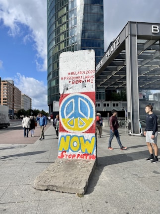 A section of the Berlin Wall with colorful graffiti depicting a peace sign and messages supporting Belarus. The background includes a modern cityscape with high-rise buildings, pedestrians, and a sign for 'PLATZ'.