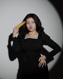 A person in a black outfit playfully holds a banana to their head against a plain backdrop, with an expression suggesting humor or playfulness.