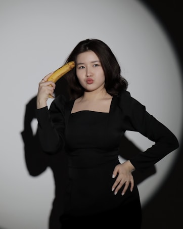 A person in a black outfit playfully holds a banana to their head against a plain backdrop, with an expression suggesting humor or playfulness.