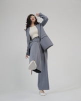 A person is shown in a dynamic pose, wearing a tailored blue-grey pantsuit and white high-heeled shoes. The background is minimalist and plain, emphasizing the fashionable nature of the outfit. The person is looking upward with a playful expression.