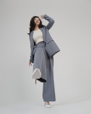 A person is shown in a dynamic pose, wearing a tailored blue-grey pantsuit and white high-heeled shoes. The background is minimalist and plain, emphasizing the fashionable nature of the outfit. The person is looking upward with a playful expression.