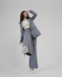 A person is shown in a dynamic pose, wearing a tailored blue-grey pantsuit and white high-heeled shoes. The background is minimalist and plain, emphasizing the fashionable nature of the outfit. The person is looking upward with a playful expression.
