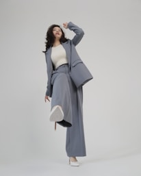 A person is shown in a dynamic pose, wearing a tailored blue-grey pantsuit and white high-heeled shoes. The background is minimalist and plain, emphasizing the fashionable nature of the outfit. The person is looking upward with a playful expression.