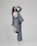A person is shown in a dynamic pose, wearing a tailored blue-grey pantsuit and white high-heeled shoes. The background is minimalist and plain, emphasizing the fashionable nature of the outfit. The person is looking upward with a playful expression.