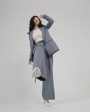 A person is shown in a dynamic pose, wearing a tailored blue-grey pantsuit and white high-heeled shoes. The background is minimalist and plain, emphasizing the fashionable nature of the outfit. The person is looking upward with a playful expression.