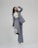 A person is shown in a dynamic pose, wearing a tailored blue-grey pantsuit and white high-heeled shoes. The background is minimalist and plain, emphasizing the fashionable nature of the outfit. The person is looking upward with a playful expression.