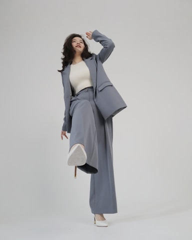 A person is shown in a dynamic pose, wearing a tailored blue-grey pantsuit and white high-heeled shoes. The background is minimalist and plain, emphasizing the fashionable nature of the outfit. The person is looking upward with a playful expression.