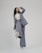 A person is shown in a dynamic pose, wearing a tailored blue-grey pantsuit and white high-heeled shoes. The background is minimalist and plain, emphasizing the fashionable nature of the outfit. The person is looking upward with a playful expression.
