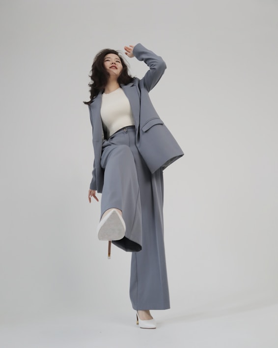 A person is shown in a dynamic pose, wearing a tailored blue-grey pantsuit and white high-heeled shoes. The background is minimalist and plain, emphasizing the fashionable nature of the outfit. The person is looking upward with a playful expression.