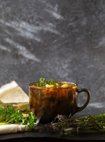 A rustic brown mug filled with a warm soup garnished with fresh herbs sits on a dark surface. Green leafy herbs are scattered around the mug, creating a natural and cozy setting. A folded white cloth adds a soft touch in the background.