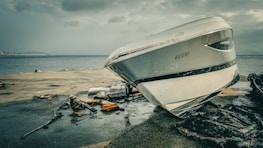A damaged boat with the name 'ECEM' is stranded on a concrete surface near the edge of a body of water. Debris and pieces of wreckage are scattered around it, possibly from the boat or other structures. The sky is overcast, adding a gloomy atmosphere to the scene.