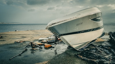 A damaged boat with the name 'ECEM' is stranded on a concrete surface near the edge of a body of water. Debris and pieces of wreckage are scattered around it, possibly from the boat or other structures. The sky is overcast, adding a gloomy atmosphere to the scene.
